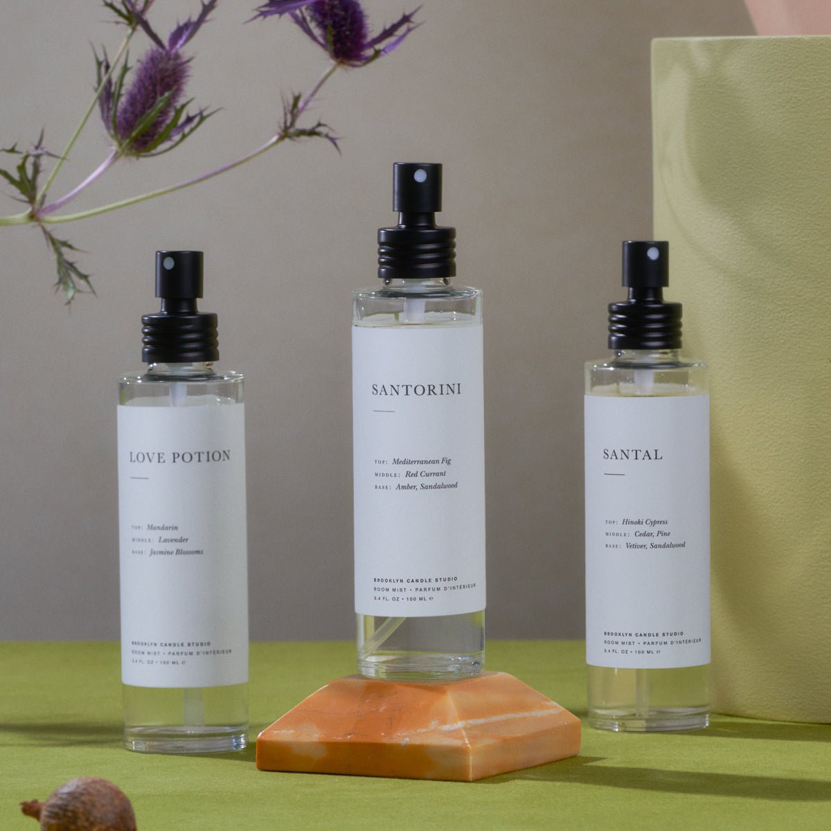 Bottles of room sprays labeled Love Potion, Santorini, and Santal on a green surface with flowers and a peach stone.