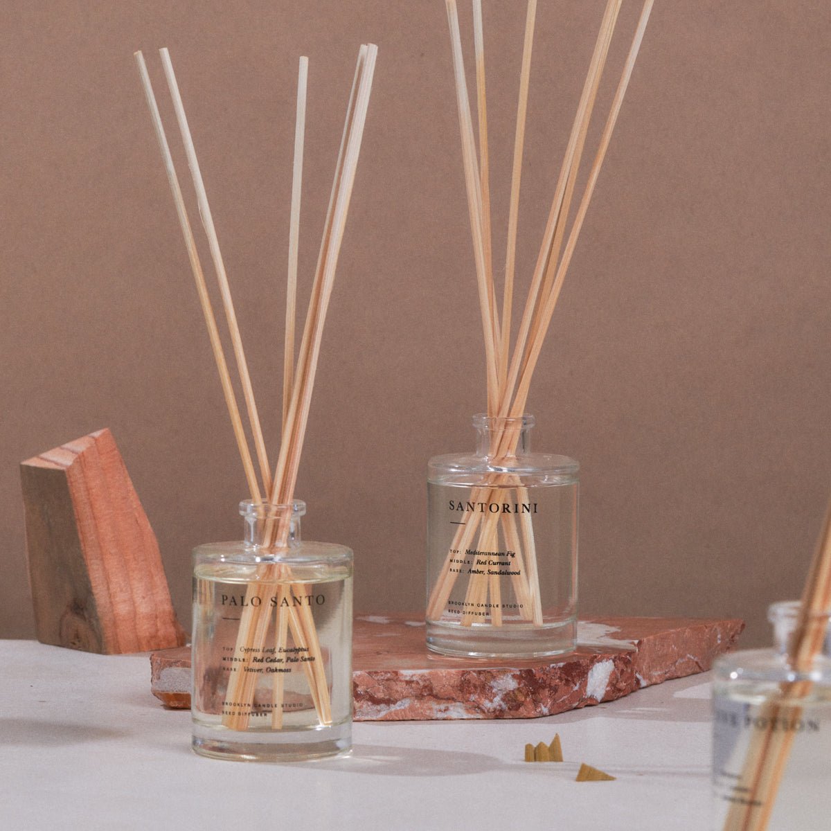 Two glass reed diffusers with wooden sticks labeled Santorini and Palo Santo on a marble and wooden surface.
